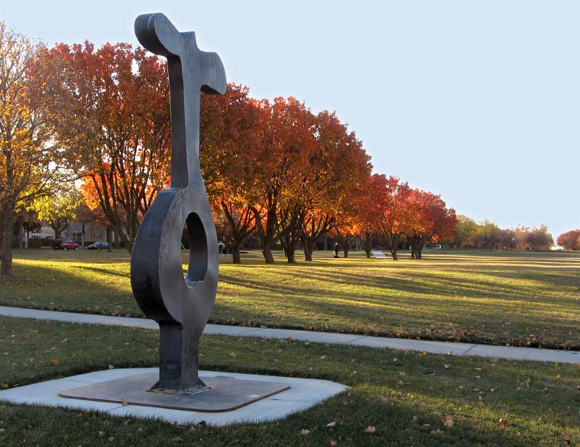 A vaguely key-like sculpture rising from the ground in front of a large grass area and trees in the fall.