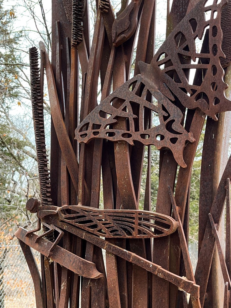 On one of the three entry panels to Pioneers Park Nature Center are some small metal bugs including a butterfly and a dragonfly in the reeds of grass. 