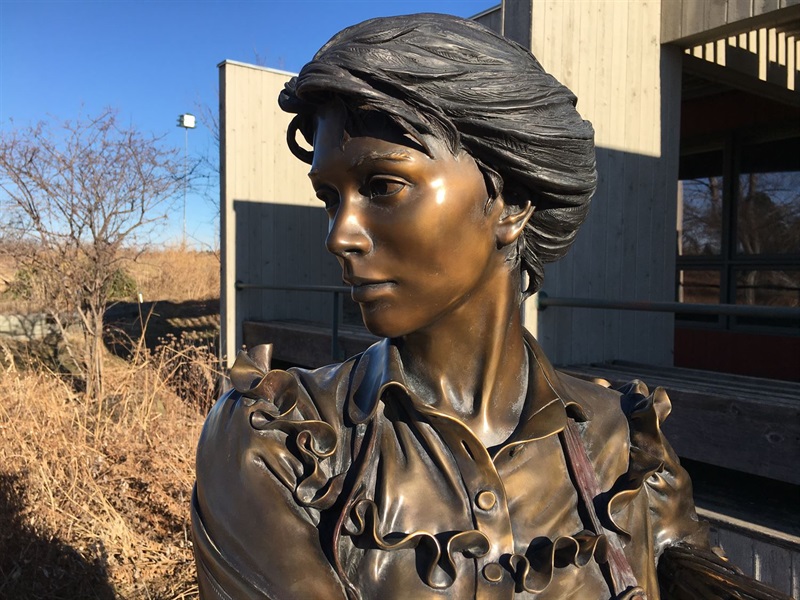 a close up of the head of the bronze sculpture of a young woman. She stands in front of a building and prairie grasses.