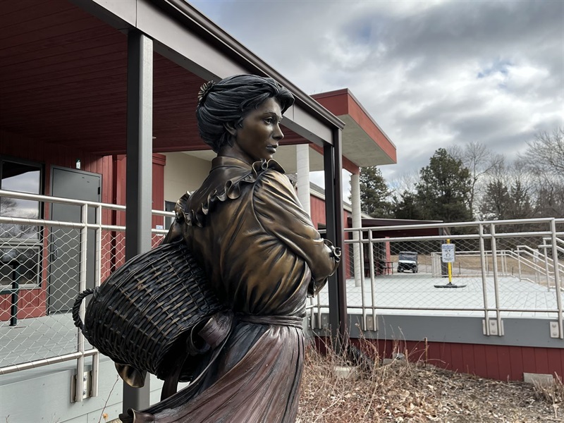 A bronze sculpture of a young woman with a basket on her hip, looking out past the building she stands in front of 