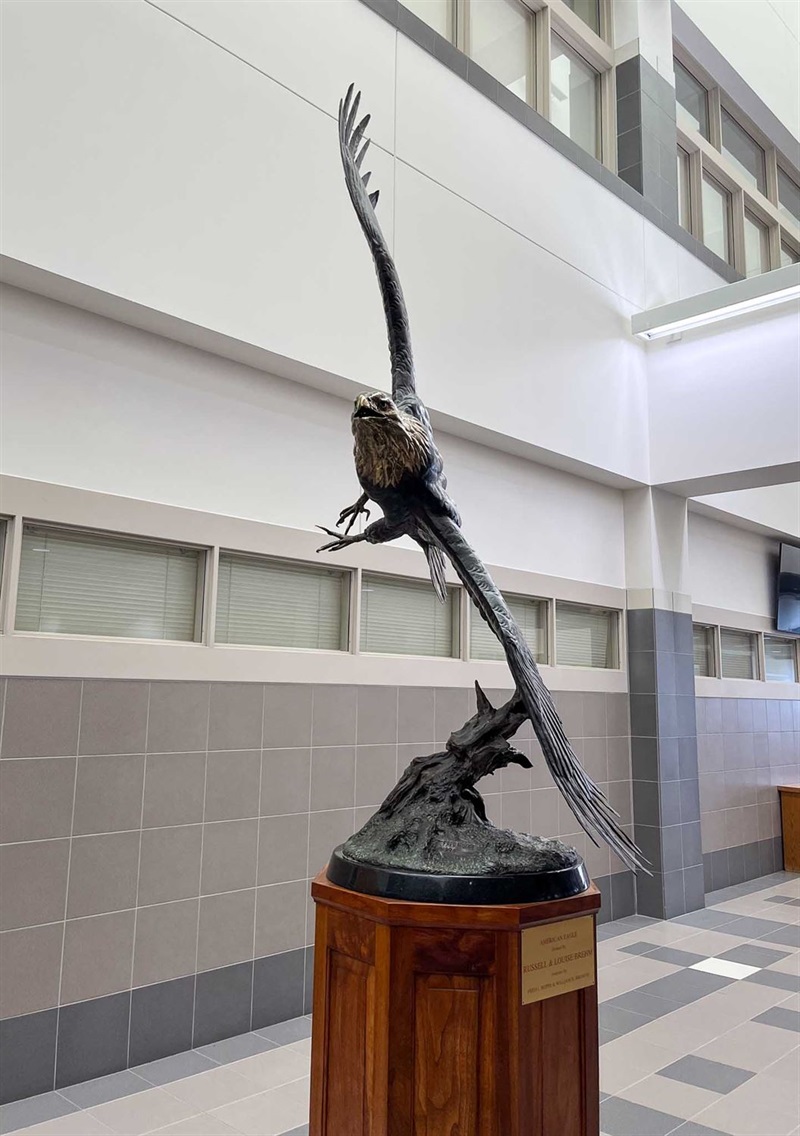 A sculpture of an eagle made of metal. The eagle is in flight with the wingspan almost entirely vertical. The eagle is hovering over a piece of wood sticking out of a rocky base, also made of metal. 