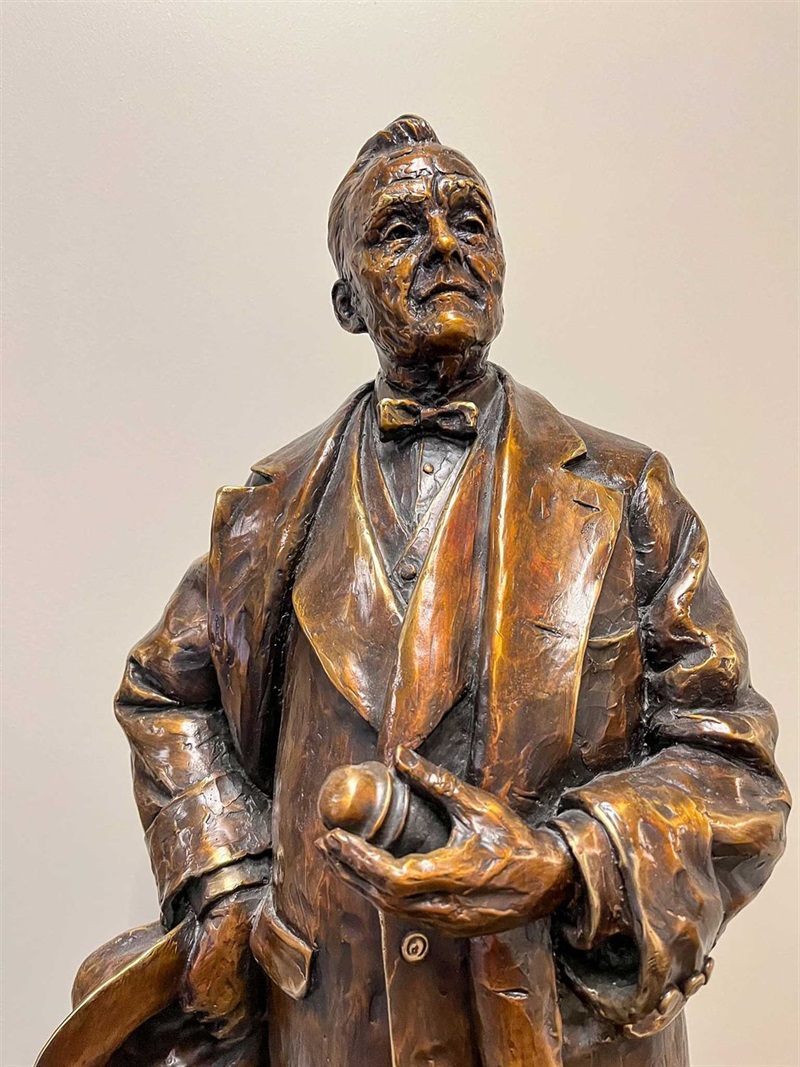 The top half of a bronze figurine of Senator George Norris. He is dressed in a suit, with a jacket and a bowtie. He is holding a glass insulator in one hand and a hat in the hand. 