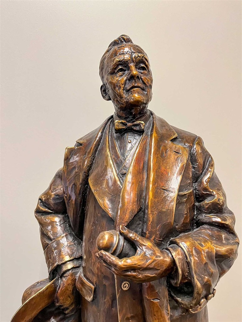 The top half of a bronze figurine of Senator George Norris. He is dressed in a suit, with a jacket and a bowtie. He is holding a glass insulator in one hand and a hat in the hand.