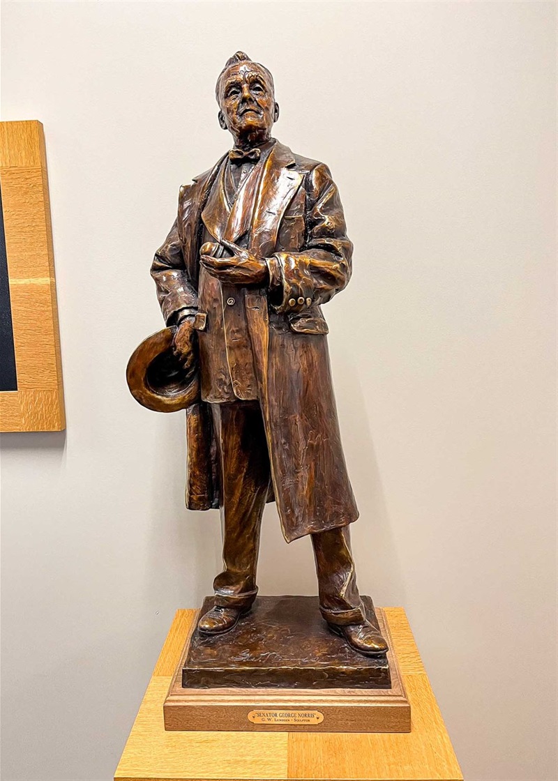 The full body of the bronze figure of George Norris. He stands with dignity, holding a bit of glass and a hat in either hand. 