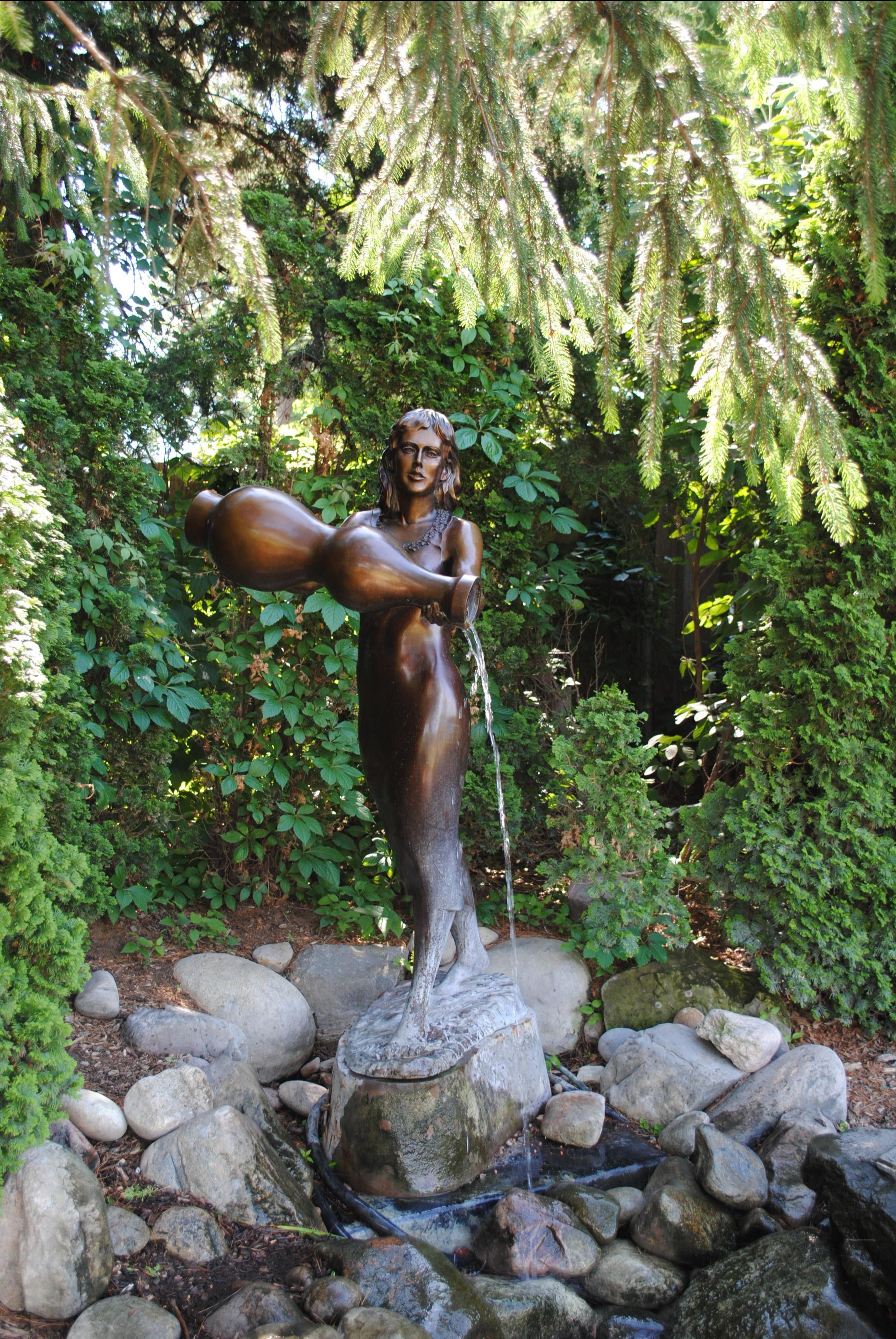 A full body shot of the bronze sculpture of Rebekah at the Well, featuring the young woman pouring the water into the water and rocks below her pedestal
