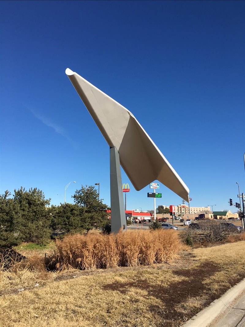 A giant paper airplane made of metal on the side of a road.