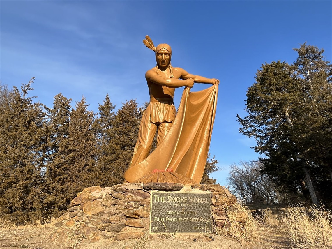 A larger than life size sculpture of an indigenous man fanning a fire with a blanket to send up a smoke signal. The sculpture is made of a brown stone.