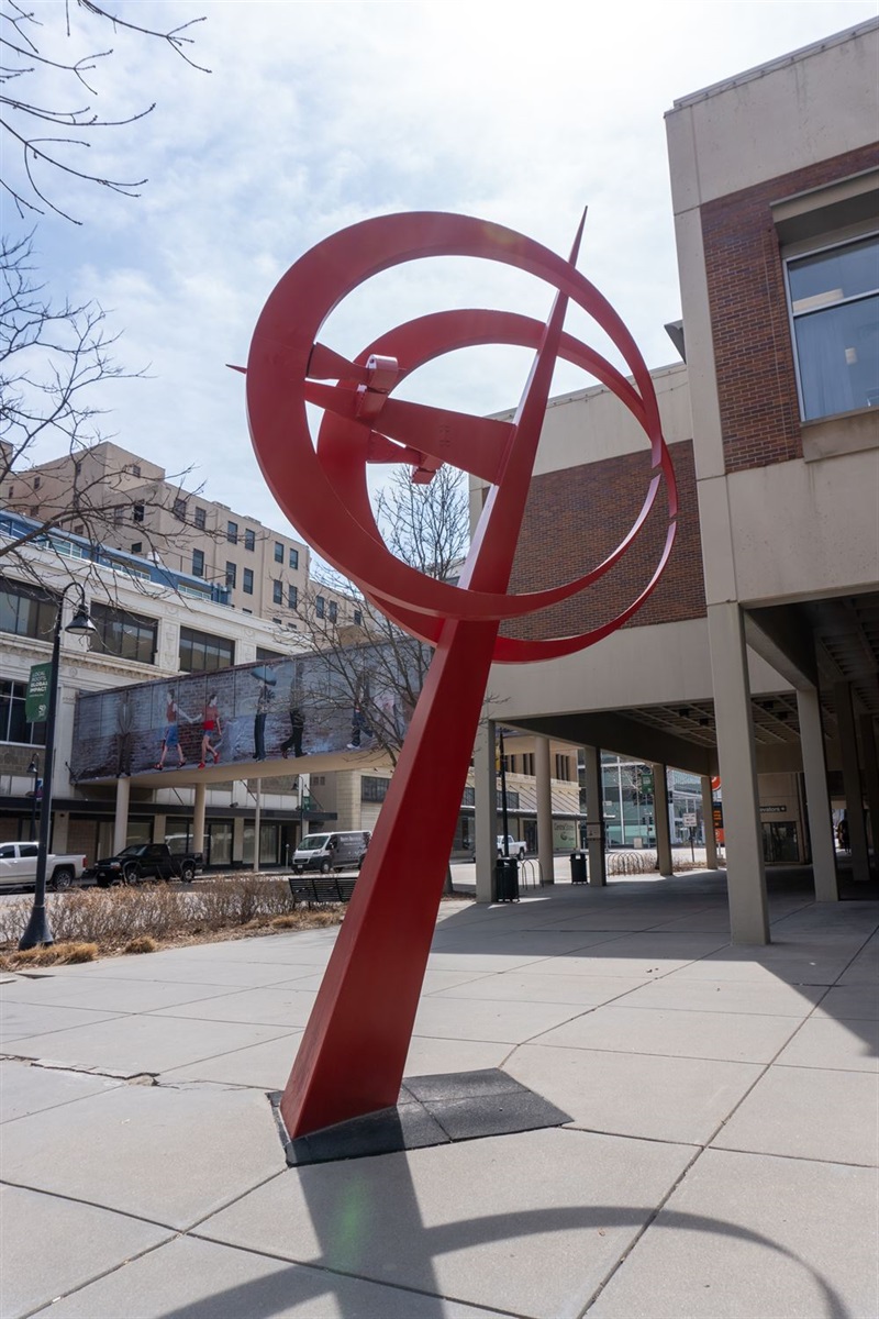 A bright red, geometric sculpture standing on the corner of a downtown street. Two red circles overlap on top of a spike