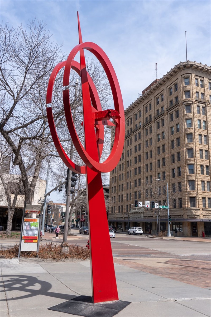 A red geometric sculpture ands on the corner of a downtown street. A large spike stands in the middle with two red circles on either side.