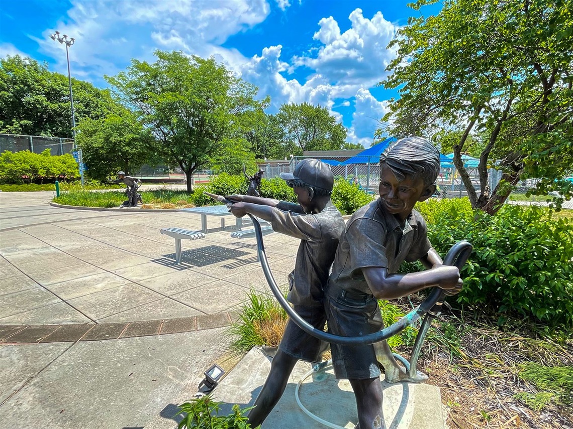 A sculpture of two young boys stand in the foreground, one spraying a hose and the other putting a kink in the rope. More kids in the water fight stand in the background.