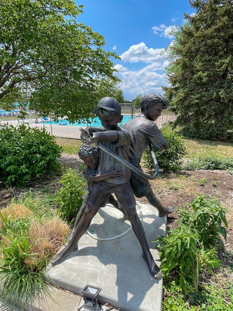 Three boys participate in a water fight with more kids we cannot see in this metal sculpture. One boys points the hose while another holds it in a kink. The third boy hides behind the two others. 