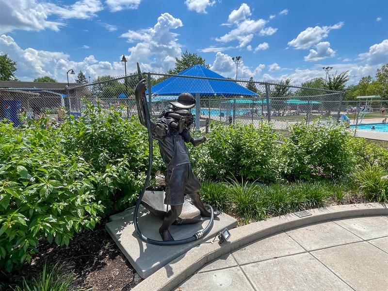 A metal sculpture of two kids playing with a water hose. The older kid holds the other on their back as well as holding an umbrella below them and the hose above them. 