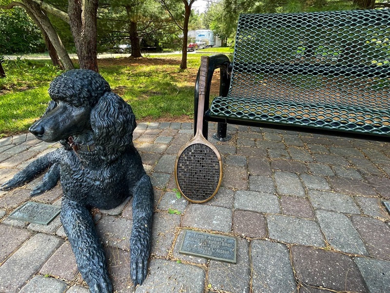 A bronze tennis racket leaning up against a park bench with a metal dog laying near it. 