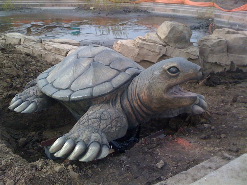 A bronze replica of turtle, laying in dirt with rocks and water behind it