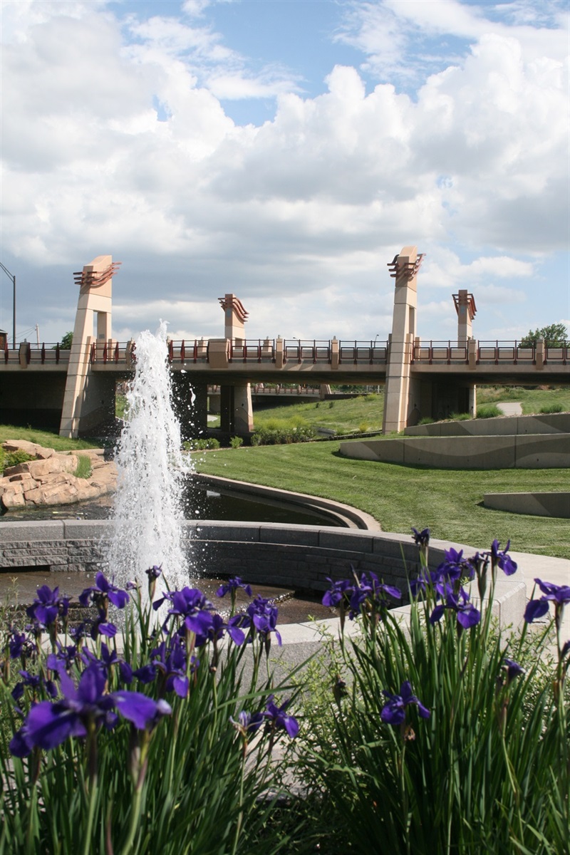 In the foreground purple flowers rise in front of the water rising from a fountain and a bridge and grass stands in the background.
