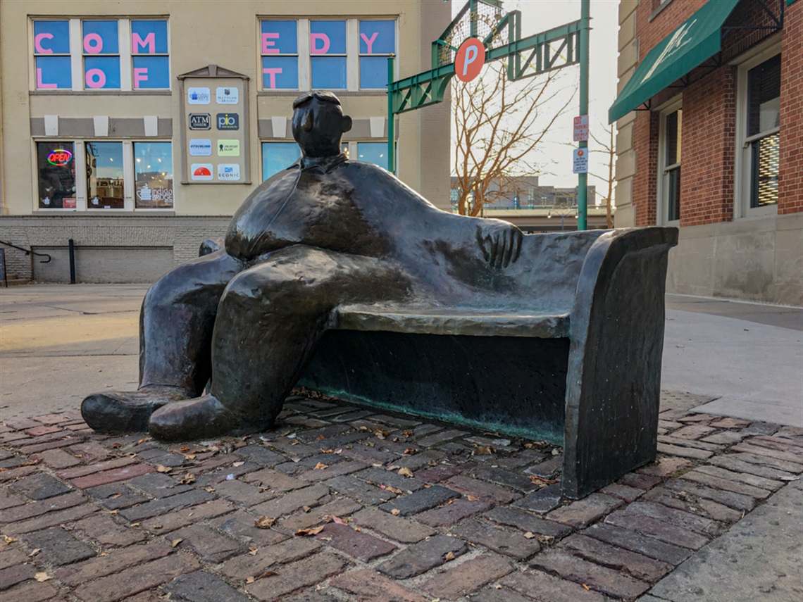 A metal bench with a metal man sitting on one side of it while looking around his surroundings. 