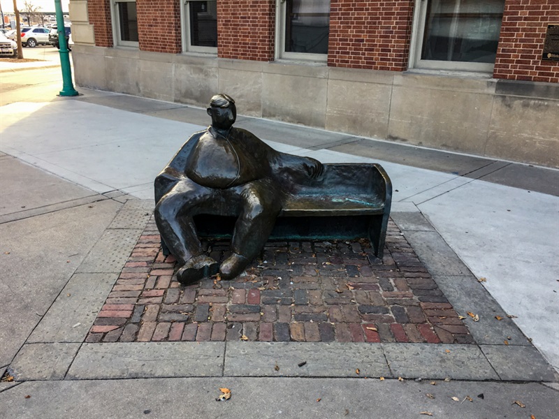 A metal bench in an urban environment, with a metal man sitting on one side of it while looking around his surroundings. 
