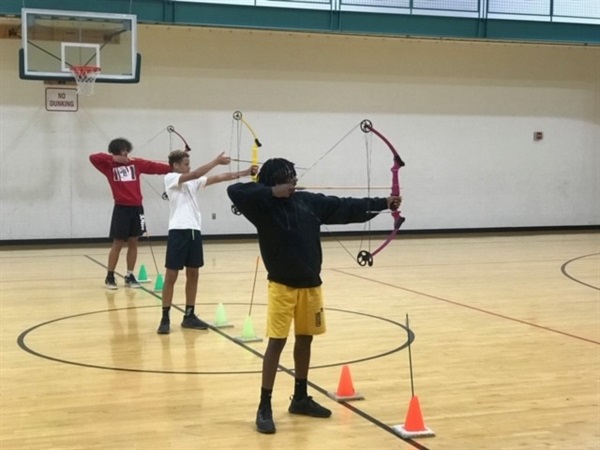 Three campers are lined up in the gym practicing archery. They're standing at half court with bows up and drawn.