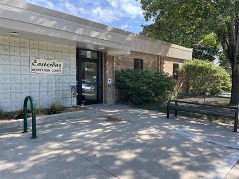The exterior of the Easterday Rec center, with a sign that says the name of the building, some bushes and trees, and a bench to sit on the sidewalk to the door. 