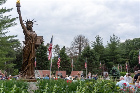 During an event at the Veterans Memorial Garden so many people stand or sit talking to each other with the Little Sister of Liberty statue in the foreground. There are American Flags posted around the garden.