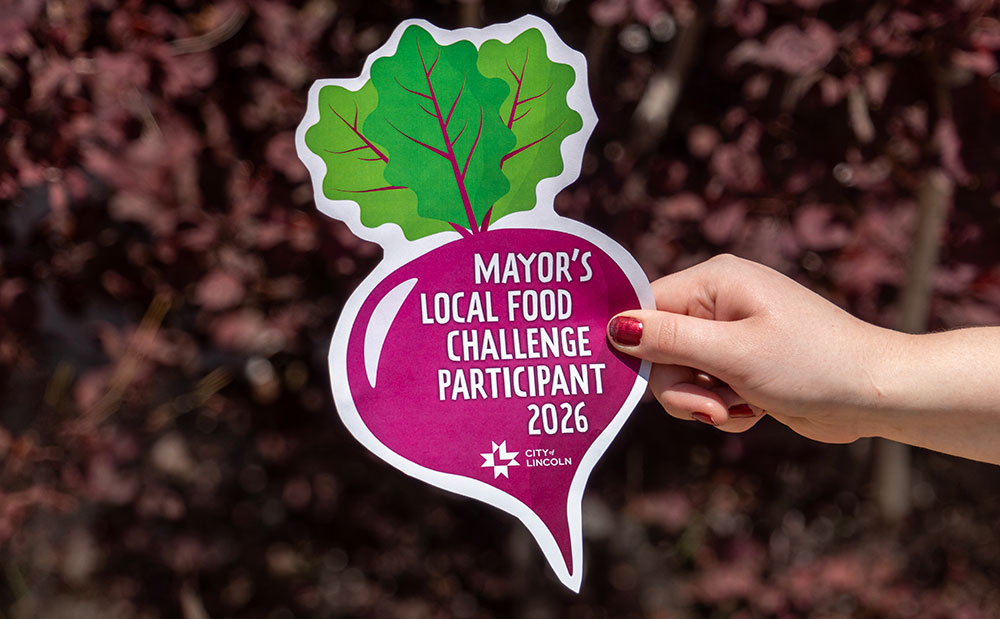 A hand holds a cut-out paper illustration of a radish with the text Mayor's Local Food Challenge Participant 2026