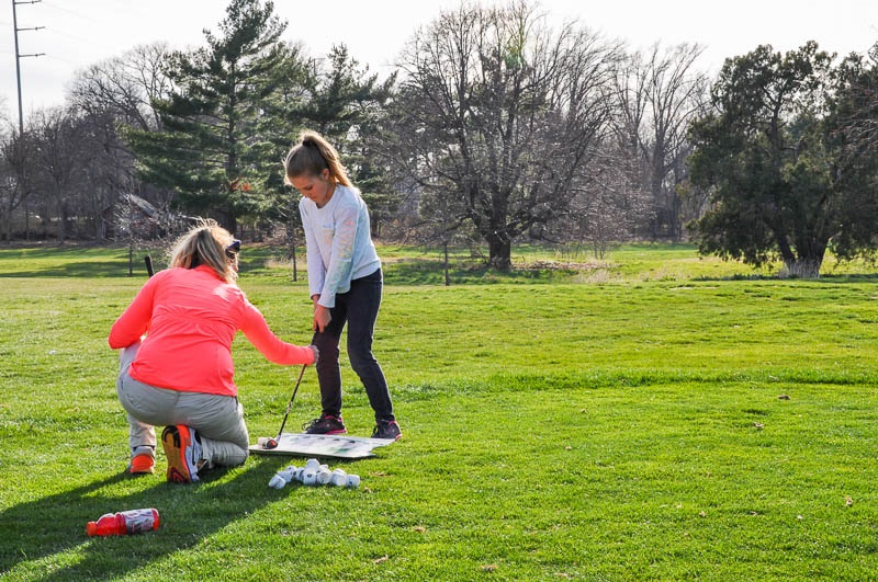 A golf instructor kneels to adjust the club of a young girl during lessons.