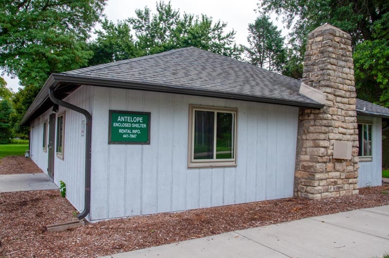 A profile shot of Antelope Enclosed shelter looking at a wall with a chimney and a Green Sign with text that reads: Antelope Enclosed Shelter Rental Info. 441-7847. 