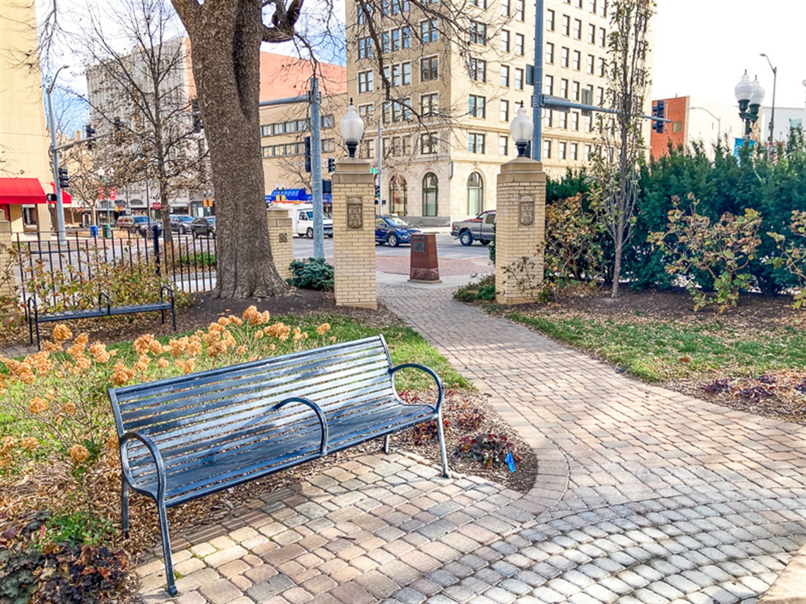 In Government Square park there is a brick paved plaza with iron benches. Behind the bench are dried hydrangea flowers in late fall flower beds, and the brick light posts marking the park’s entrance.