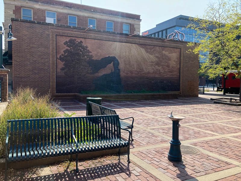 A brick paved plaza with benches native grasses, and a 3-D brick mural of Locomotive 710.
