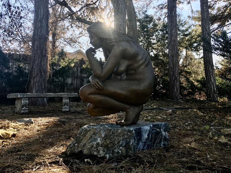 A metal figure of a woman kneeling, looking towards the ground in contemplation surrounded by trees and grass