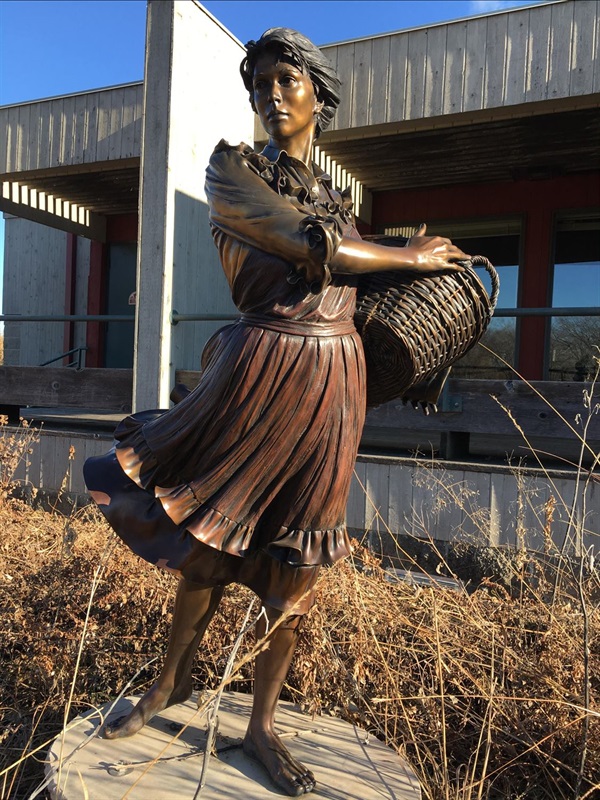 A bronze statue of a young woman with a basket on her hip, surrounded by grasses