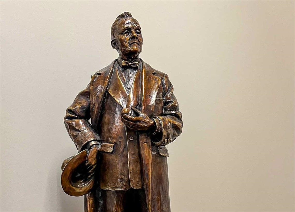 The top half of a bronze figure of Senator George Norris. He is holding a bit of glass in one hand and a hat in the other. He looks outward with dignity. 