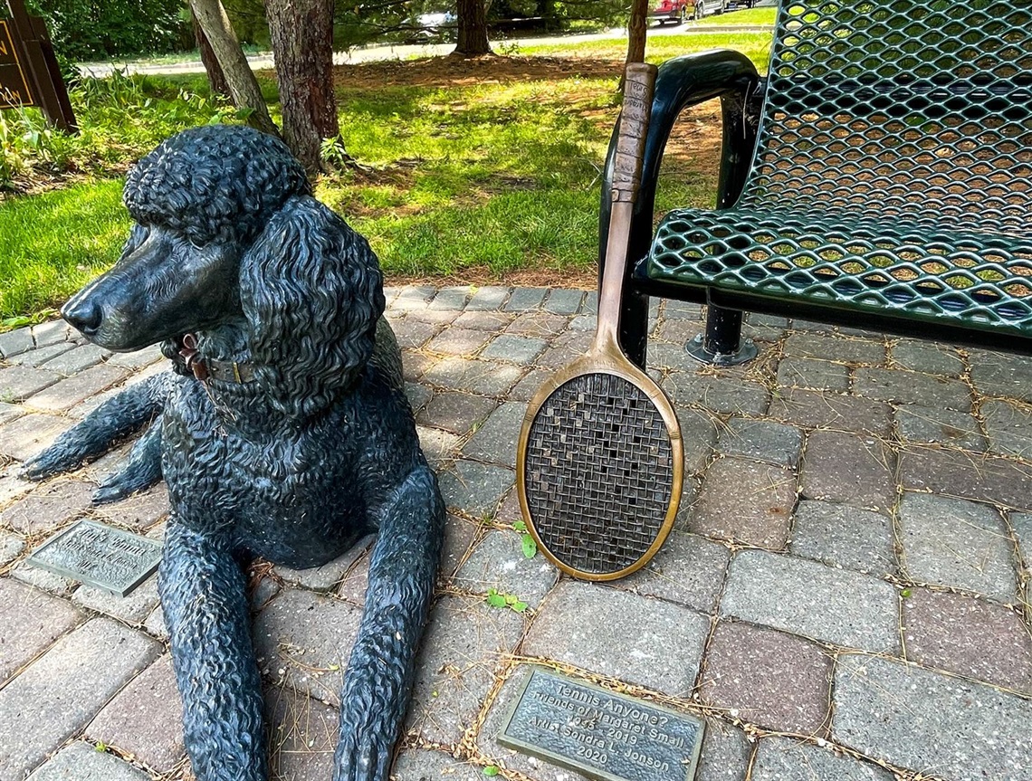 A bronze tennis racket leaning up against a park bench with a metal dog laying near it. 