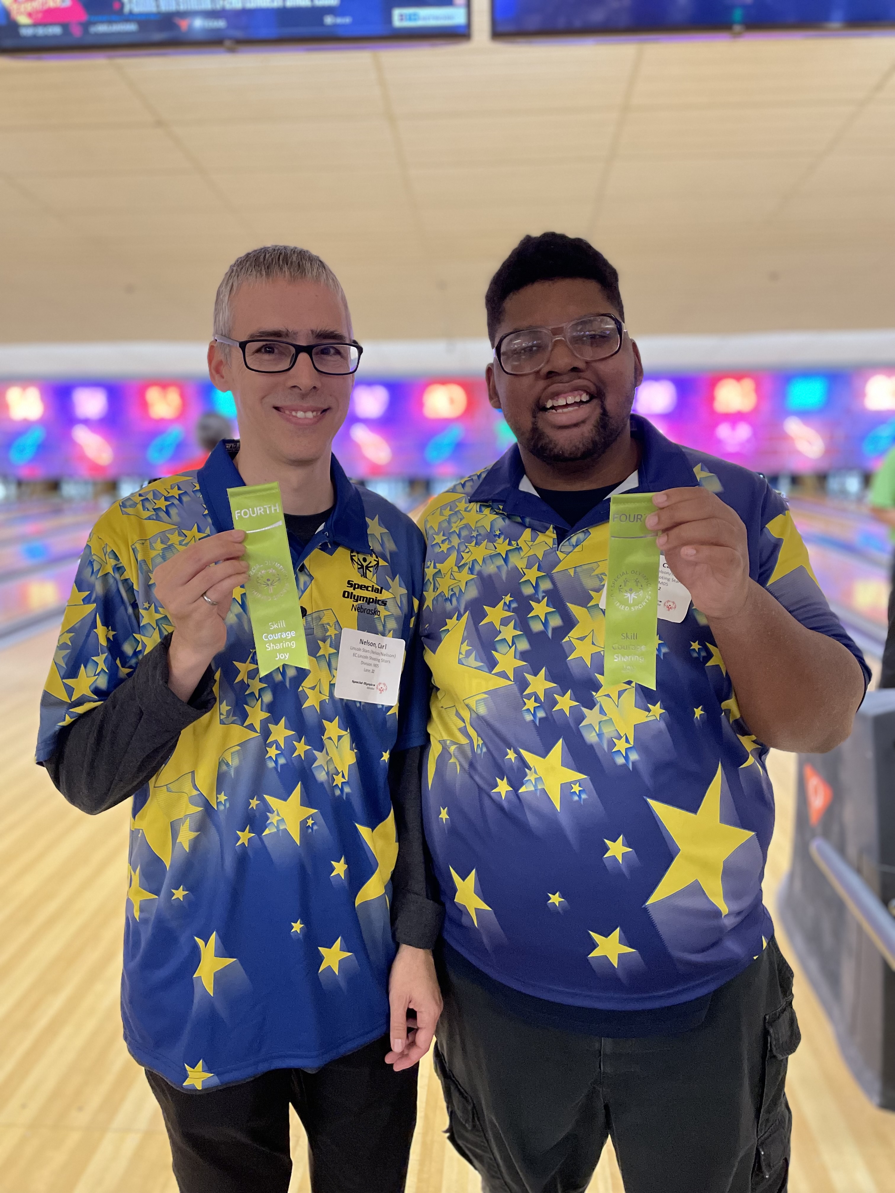 Two bowlers in uniform hold up ribbons in a bowling alley.