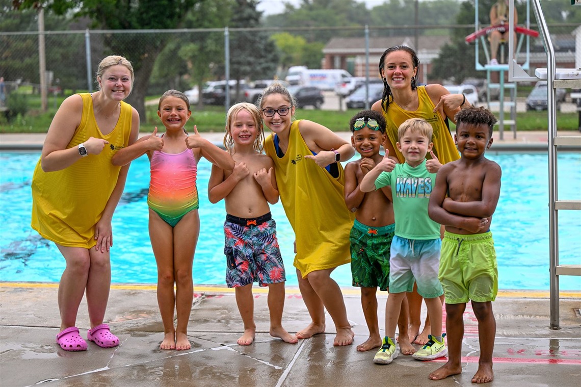 A group of children and camp leaders pose for a photo while standing on a pool deck with the pool in the background.