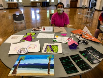 Camper shows of their Art Camp projects. A variety of paintings, prints, and sculptural projects are artistically laid out on a table in front of the camper. 
